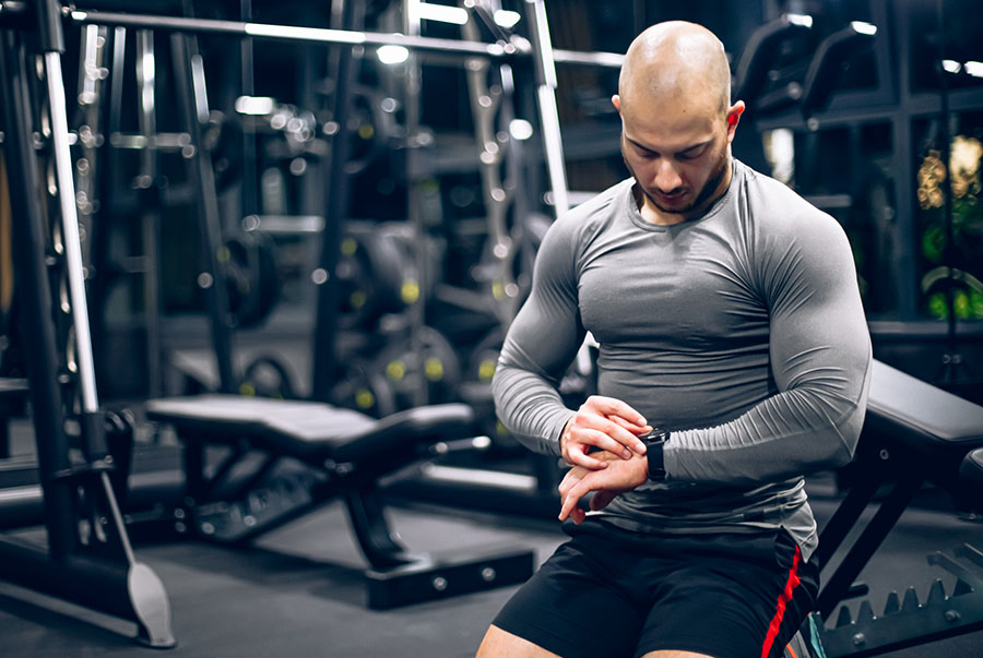 Muscular man in gym checking his watch between sets, symbolizing time-efficient strength training for busy people.